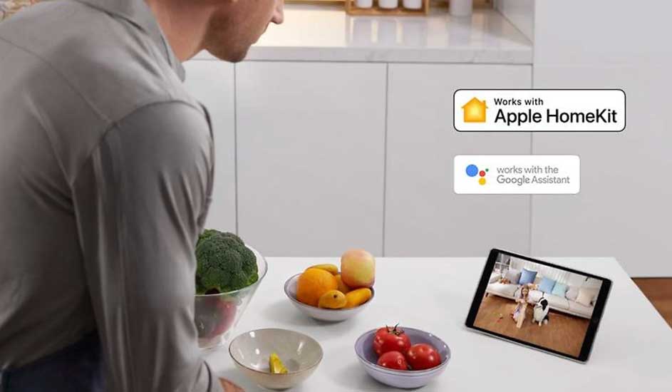 Een man staat in de keuken met fruit en groenten op het aanrecht. Op een tablet ziet hij een kind en hond. Apple HomeKit-logo zichtbaar.