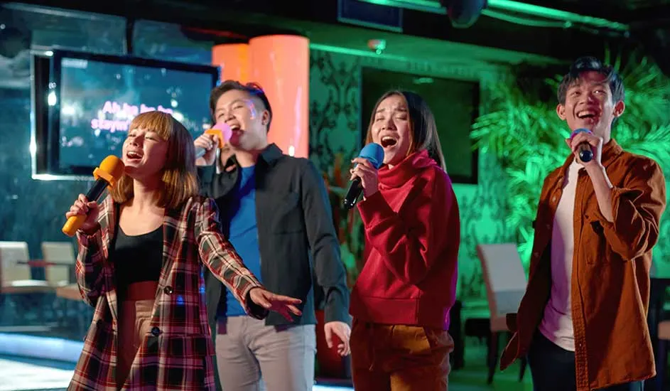 Quatre personnes chantent dans un bar karaoké, tenant des microphones de différentes couleurs. Des écrans et des lumières colorées sont visibles en arrière-plan.