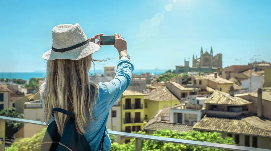 Une touriste en vacances avec un chapeau prend une photo d'une ville avec un smartphone sous un ciel bleu.