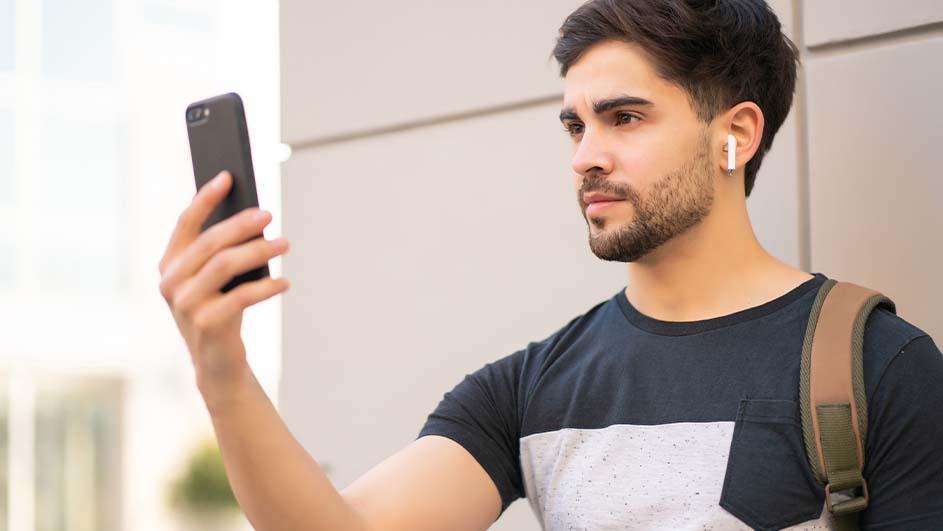 Un homme tient un smartphone noir devant un mur blanc. Il porte un t-shirt foncé et un sac à dos.