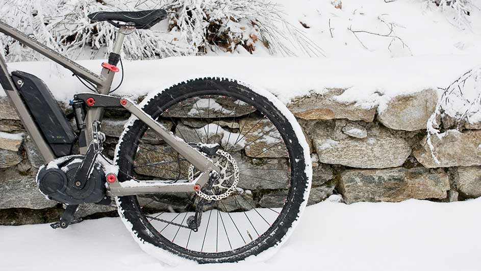 Een grijze elektrische fiets staat in de sneeuw naast een stenen muur.