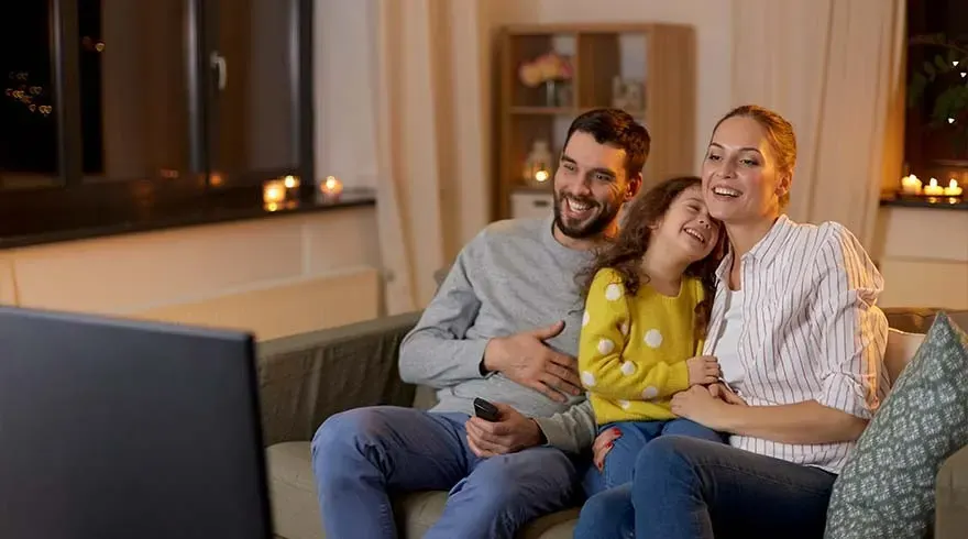 A happy family sits together on the couch watching television. A man, woman and child laugh and enjoy. Warm, cozy living room with candles and evening lighting.