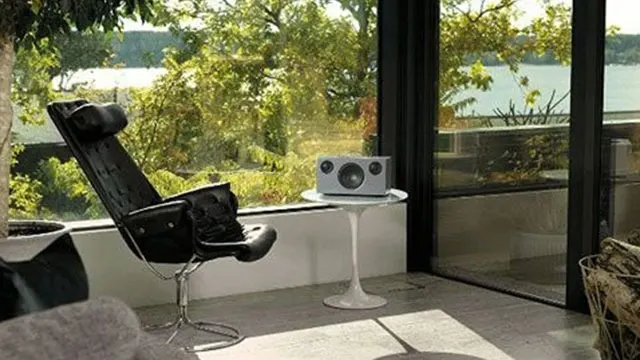 Een interieur met een zwarte stoel, een witte tafel met een grijze luidspreker, en een groot raam met uitzicht op groen en water.