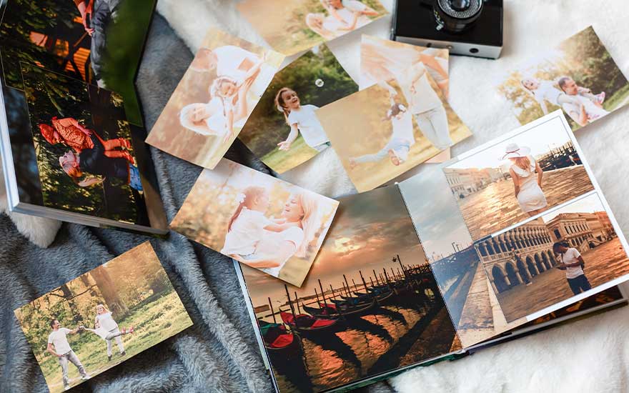 Photo album, loose prints, and a camera on a gray blanket. Prints show outdoor scenes and a Venice canal view inside the album.