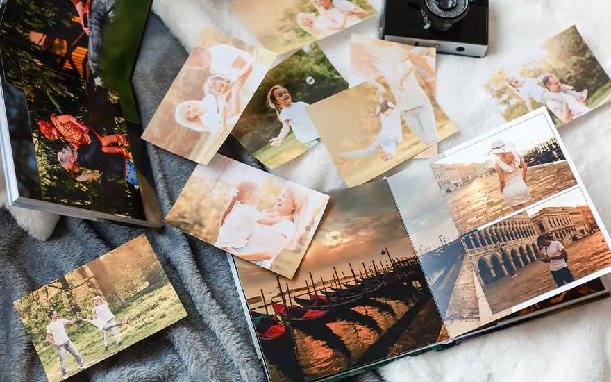 Photo album, loose prints, and a camera on a gray blanket. Prints show outdoor scenes and a Venice canal view inside the album.