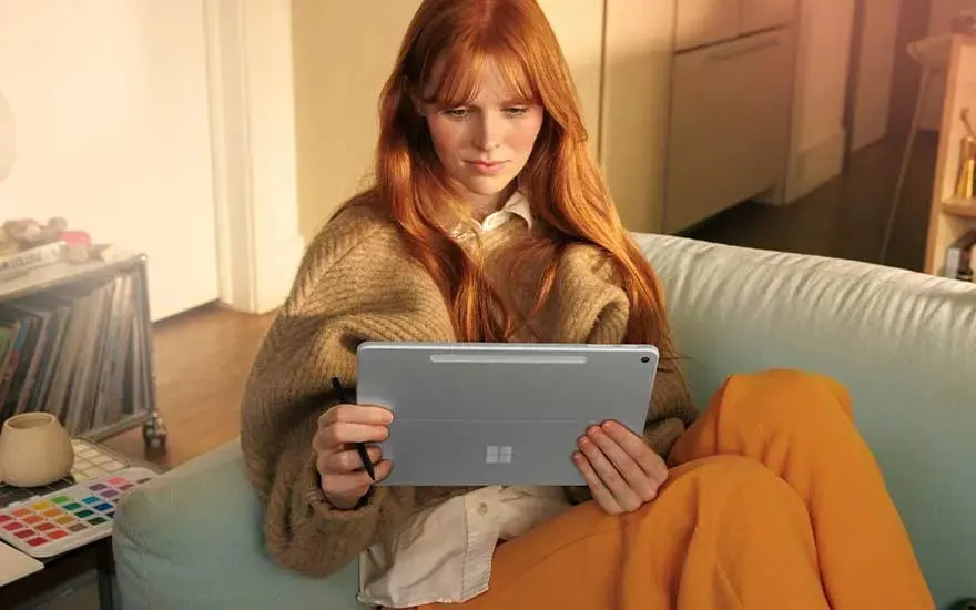 Vrouw met rood haar en bruine trui gebruikt tablet op bank, oranje dekentje over benen, warme huiselijke sfeer met planten.