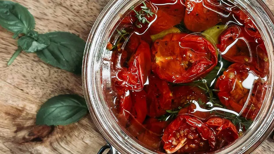 Vue de dessus d'un bocal en verre rempli de tomates séchées rouges et d'herbes, sur une table en bois avec du basilic.