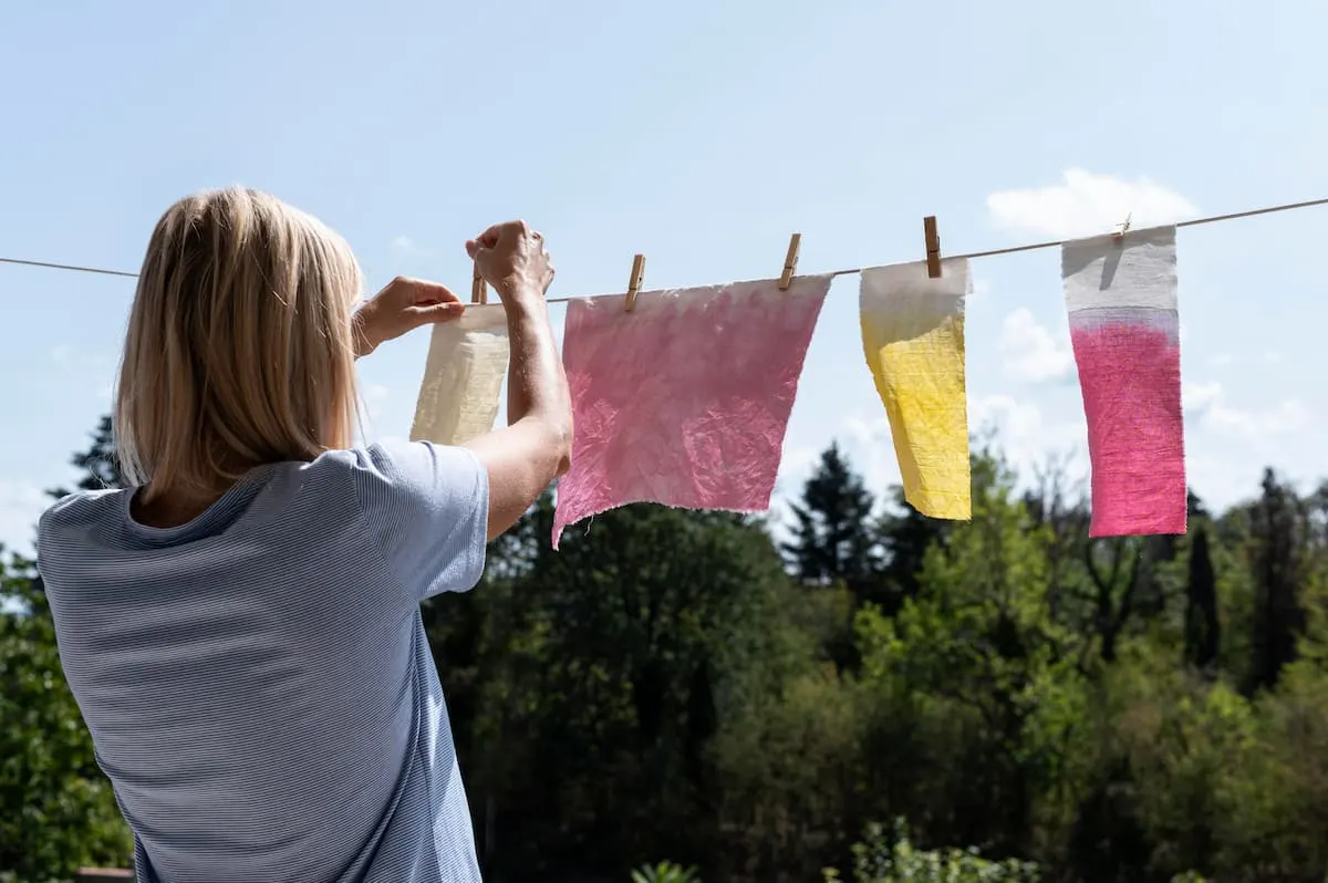 Imagen de una chica tendiendo paños de colores en una cuerda de exterior. Fondo de cielo azul con nubes blancas y un bosque debajo.