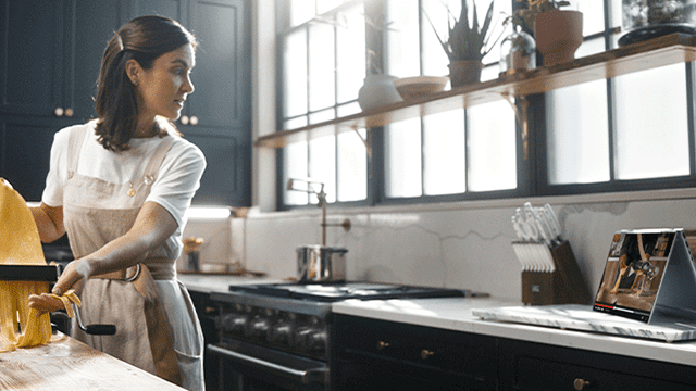 Mujer en delantal usa máquina para pasta en cocina iluminada. Horno, estantes con plantas, y laptop visibles.