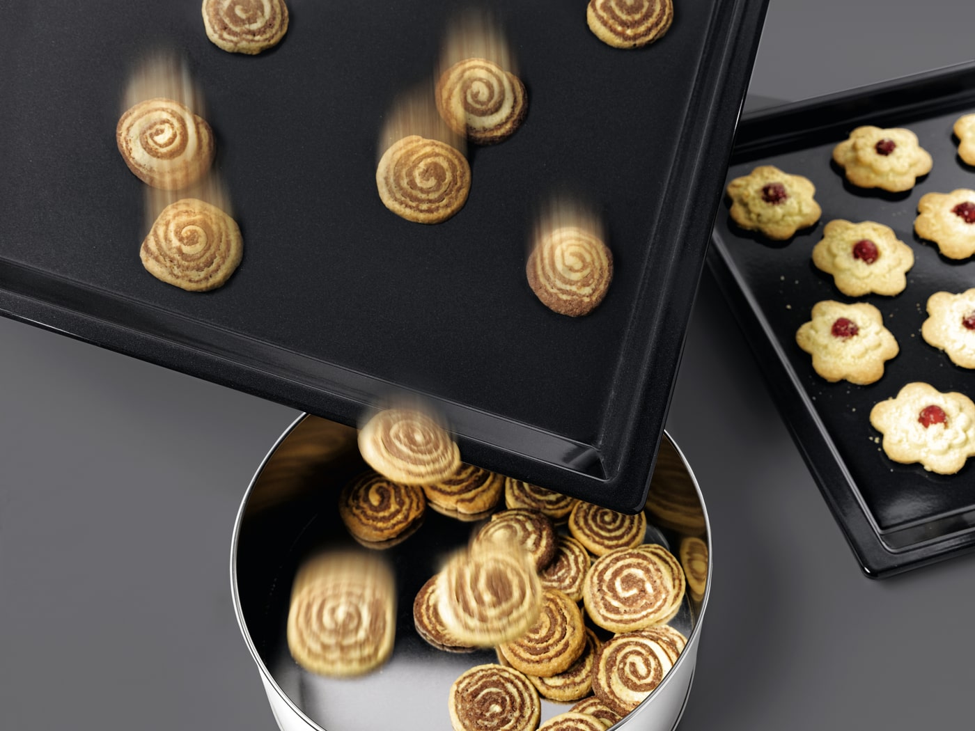 Galletas en espiral cayendo de una bandeja negra a una lata plateada. Otra bandeja con galletas de flor en segundo plano.