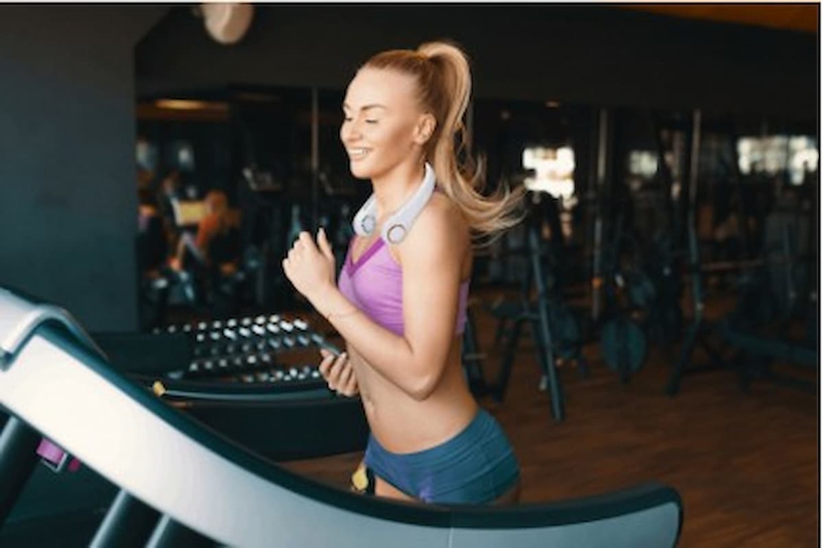 Mujer joven sonriente corriendo en una caminadora con audífonos en el cuello dentro de un gimnasio.