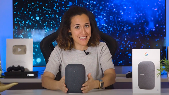 Mujer sonriente sujeta un altavoz gris de Google. Al fondo, un monitor muestra un diseño abstracto en azul y blanco.