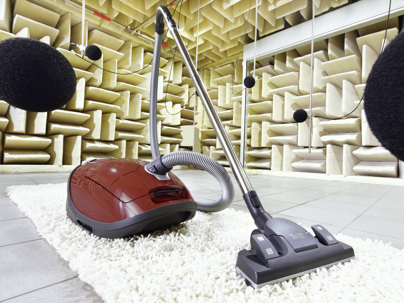Aspiradora roja y gris sobre alfombra blanca en una habitación con paneles acústicos y micrófonos colgantes.