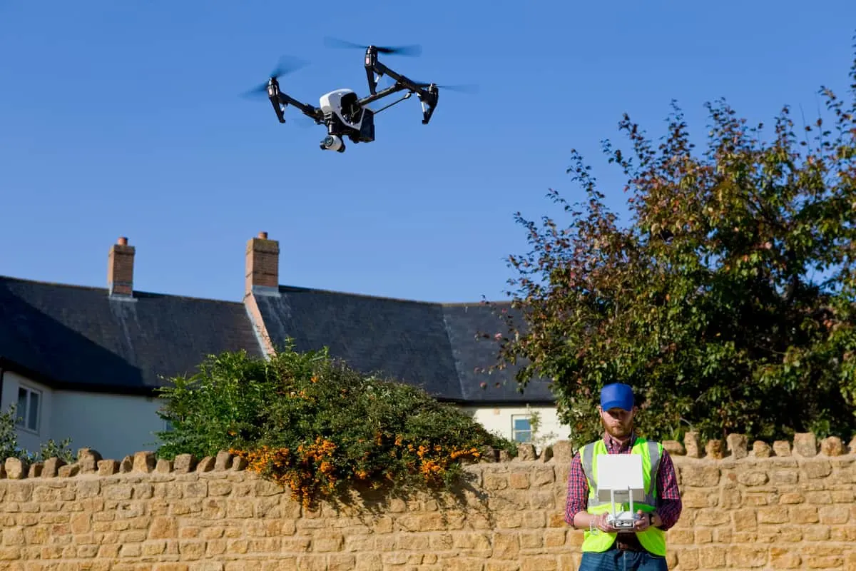 Operador con chaleco de seguridad controlando un dron en vuelo sobre un muro de piedra con casas al fondo.