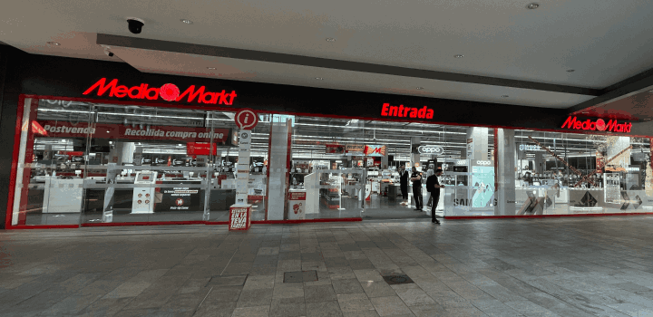Fachada de tienda MediaMarkt con letreros rojos, entrada de cristal y suelo de baldosas gris. Hay gente entrando.