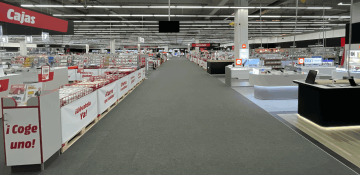 Pasillo extenso de tienda con estantes de productos y letreros rojos. Una caja registradora negra está a la derecha.