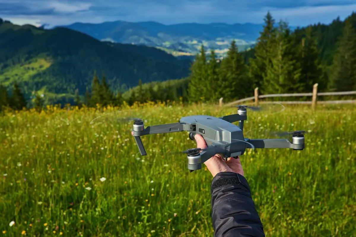 Mano sosteniendo un dron listo para despegar en un prado verde rodeado de montañas.