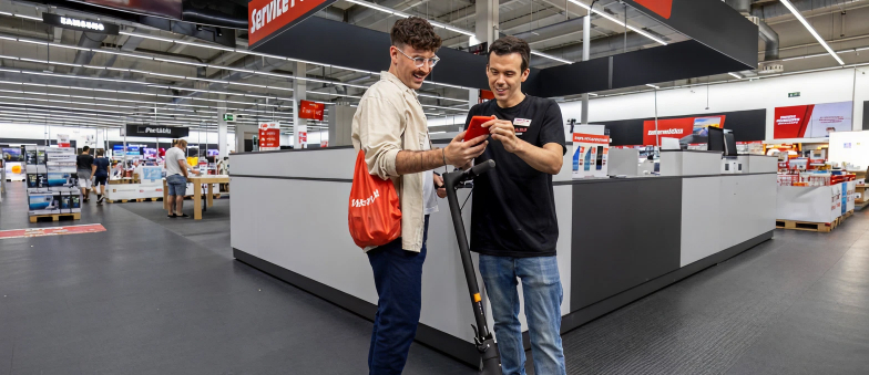 En una tienda, un hombre en patinete eléctrico mira un móvil rojo que le muestra otro hombre cerca de cajas registradoras.