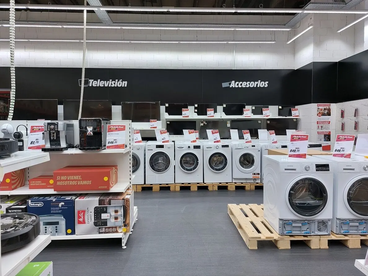 Interior de tienda con lavadoras blancas sobre palés de madera, televisores y estanterías con cajas y electrodomésticos. Señalización de "Televisión" y "Accesorios".