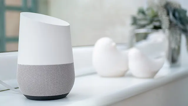 Un altavoz inteligente blanco y gris sobre una superficie blanca. Dos objetos blancos y una planta están al fondo.