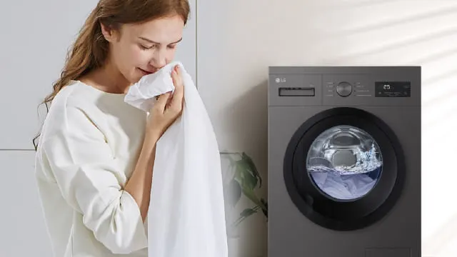 Persona disfrutando del aroma de una sábana junto a una lavadora de carga frontal gris en funcionamiento.