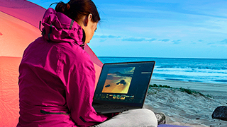 Mujer con chaqueta rosa y portátil en playa arenosa. Océano azul de fondo. Tienda naranja a la izquierda.