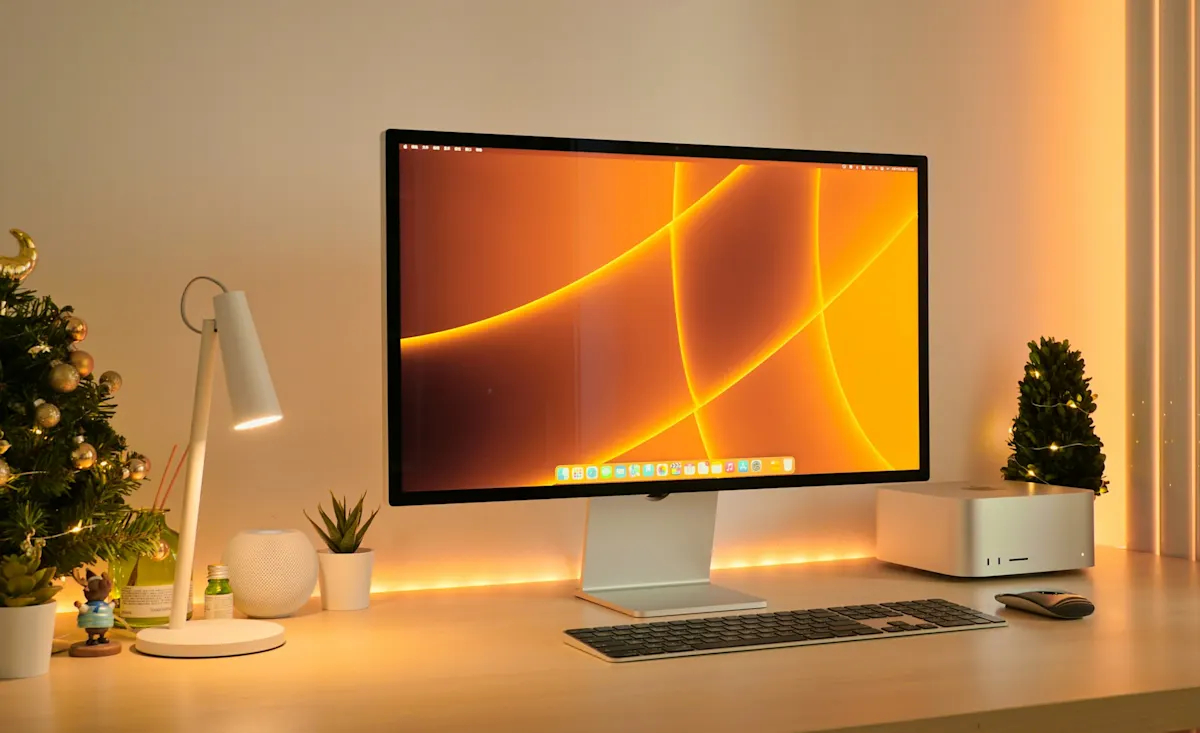 Escritorio minimalista con monitor grande iluminado en tonos cálidos, acompañado de teclado, ratón, lámpara y decoraciones navideñas.