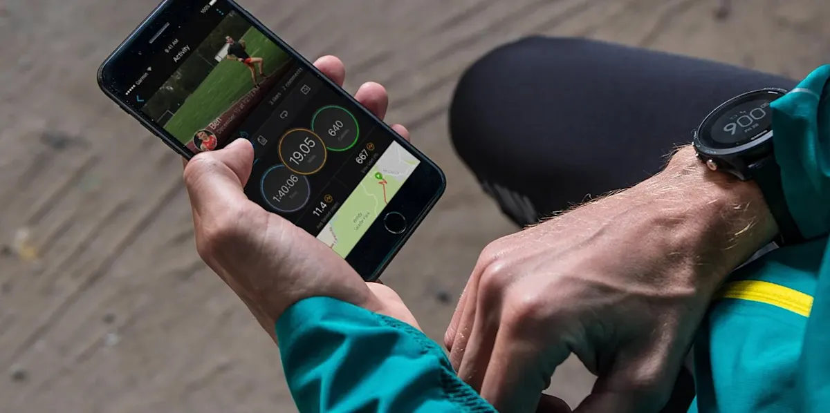 Persona revisa datos deportivos en móvil y reloj digital. El móvil muestra tiempo, distancia y mapa. Lleva chaqueta verde con franja amarilla.