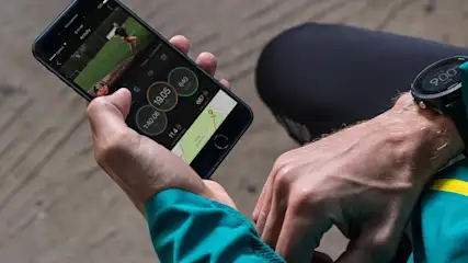 Persona revisa datos deportivos en móvil y reloj digital. El móvil muestra tiempo, distancia y mapa. Lleva chaqueta verde con franja amarilla.