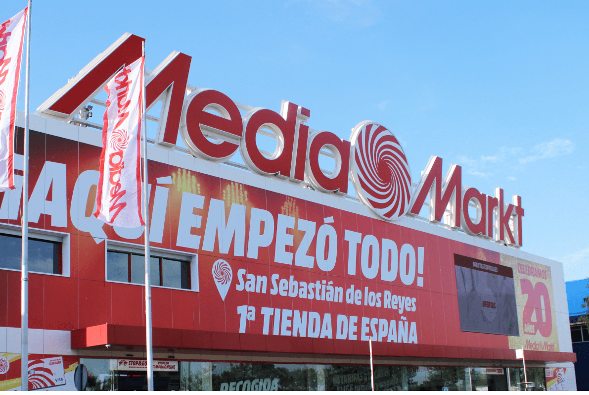 Edificio de Media Markt con letreros rojos y blancos bajo cielo azul. Banderas ondean al frente del local.