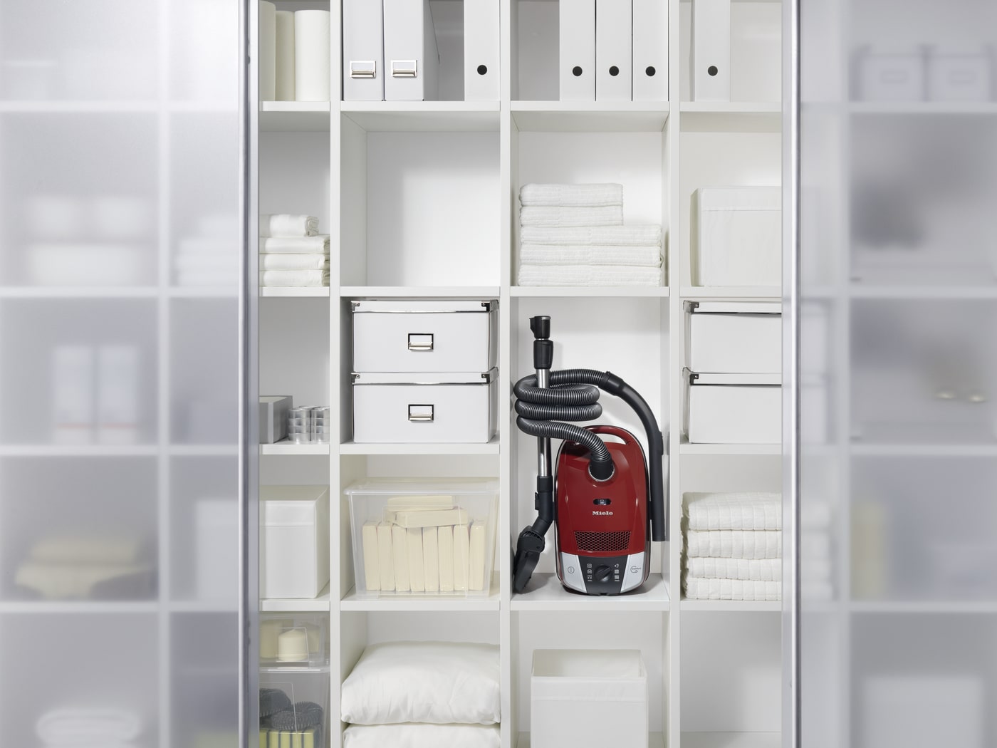 Estantería blanca con carpetas, toallas, cajas y una aspiradora roja. Puerta corredera con vidrio esmerilado a la izquierda.