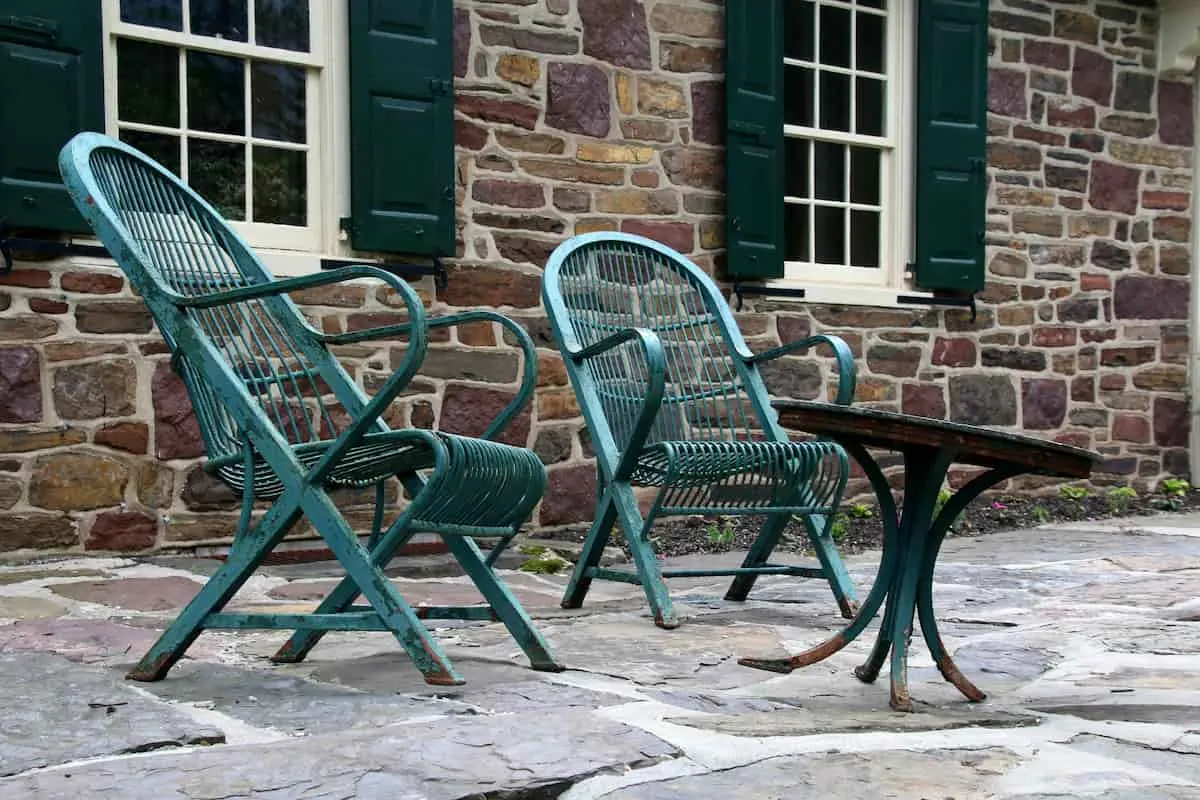 Imagen de dos sillas de jardín de metal, inclinadas hacia atrás y con reposabrazos. Delante una pequeña mesa circular también de metal con una de las patas inclinadas. Mesa y sillas de color verde turquesa. Suelo de piedra de pueblo rural con fondo de una casa rural también de piedra con dos ventanas.