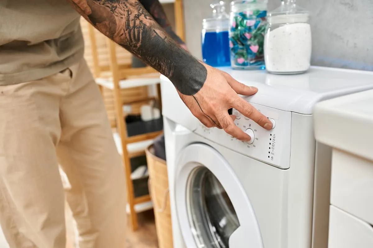 Brazo tatuado de una persona ajustando el dial de programas en la parte frontal de una lavadora blanca en una zona de lavandería con detergentes sobre la tapa.