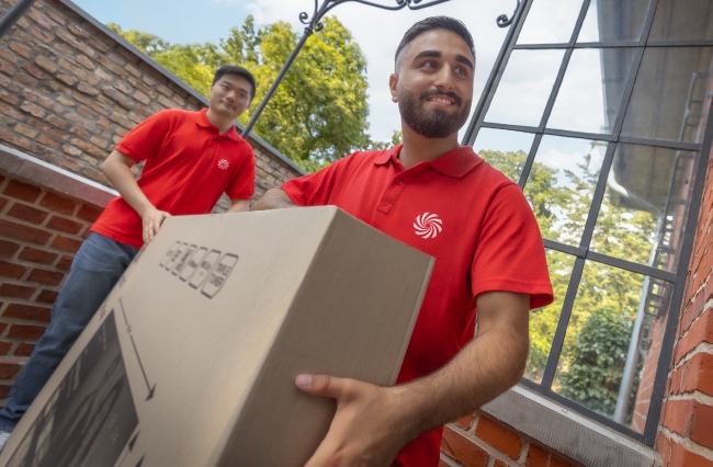 Dos hombres en camisas rojas llevan una caja grande frente a una ventana y una pared de ladrillos.
