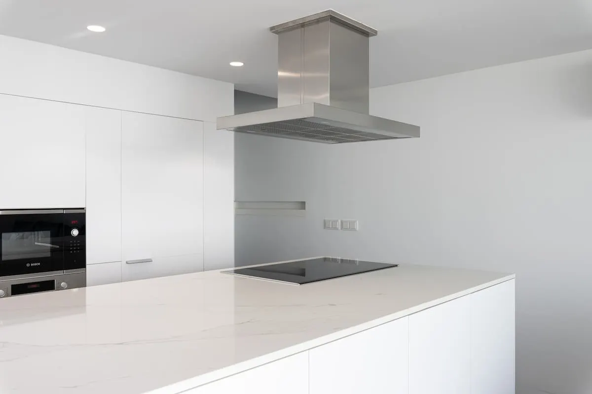 Cocina moderna minimalista blanca con isla de mármol, placa de inducción y campana extractora de acero inoxidable.