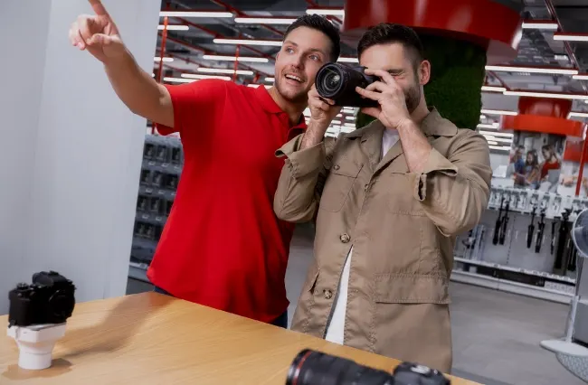 Dos hombres en una tienda de cámaras; uno vestido de rojo señala y el otro prueba una cámara.
