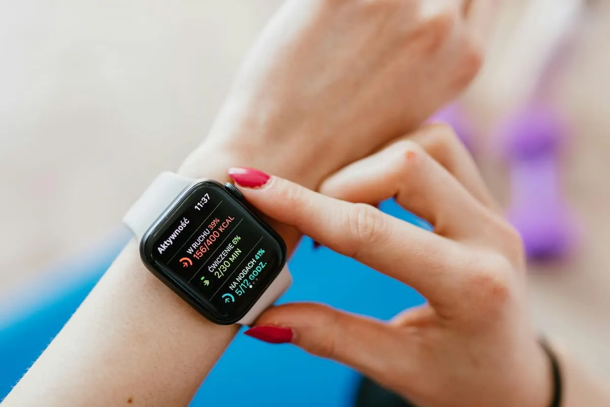 Persona con uñas pintadas de rojo usando un smartwatch con correa blanca que muestra datos de actividad física en la pantalla.