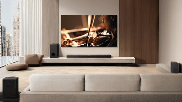 Salón con sofá beige frente a la TV que muestra violinista. Un altavoz y barra de sonido están en un estante blanco. Ventana a la izquierda.