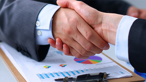 Dos personas vestidas con traje se dan la mano sobre documentos con gráficos y un portapapeles.