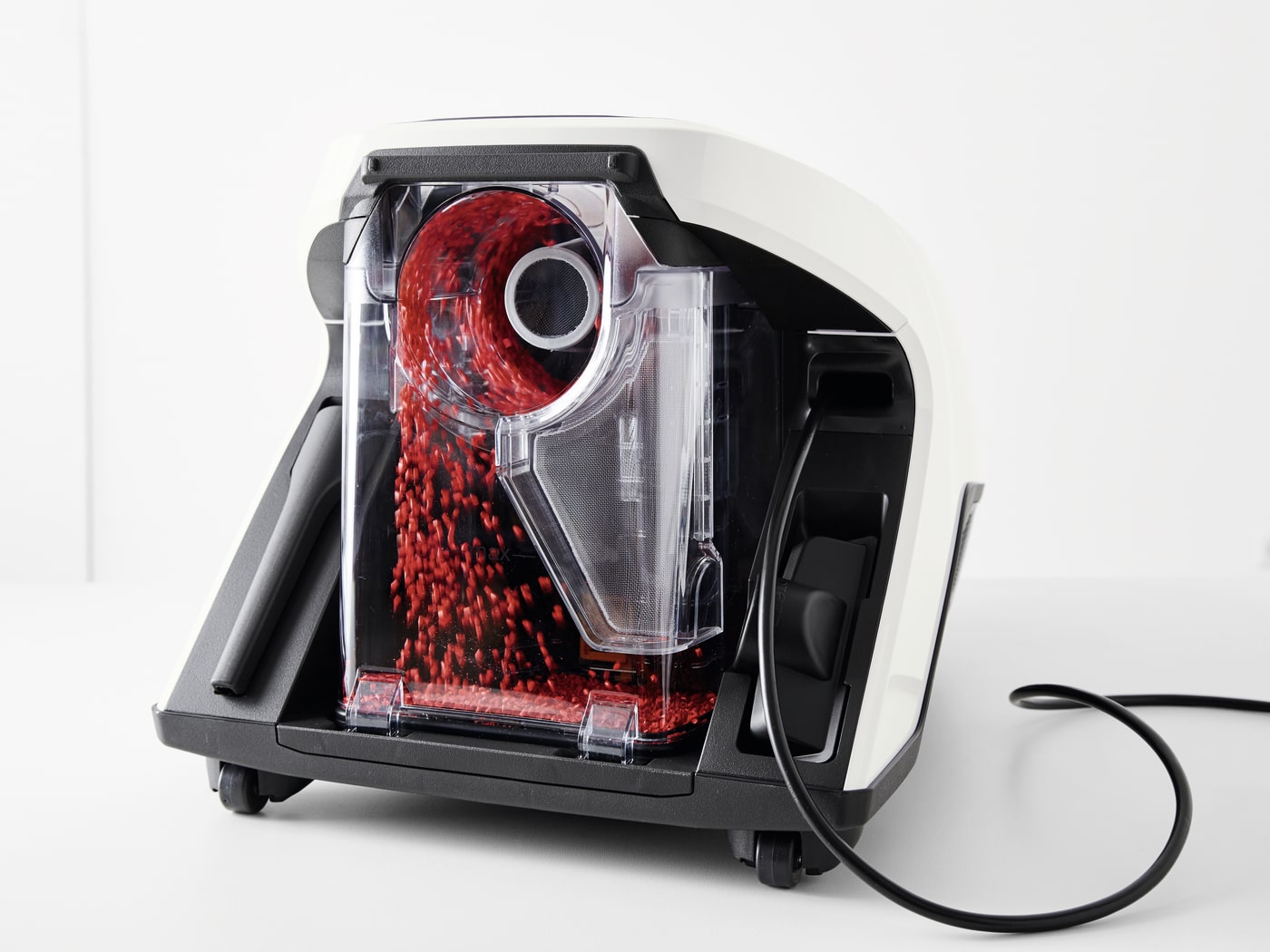 Máquina blanca y negra con recipiente transparente lleno de granos rojos, con cable eléctrico negro.