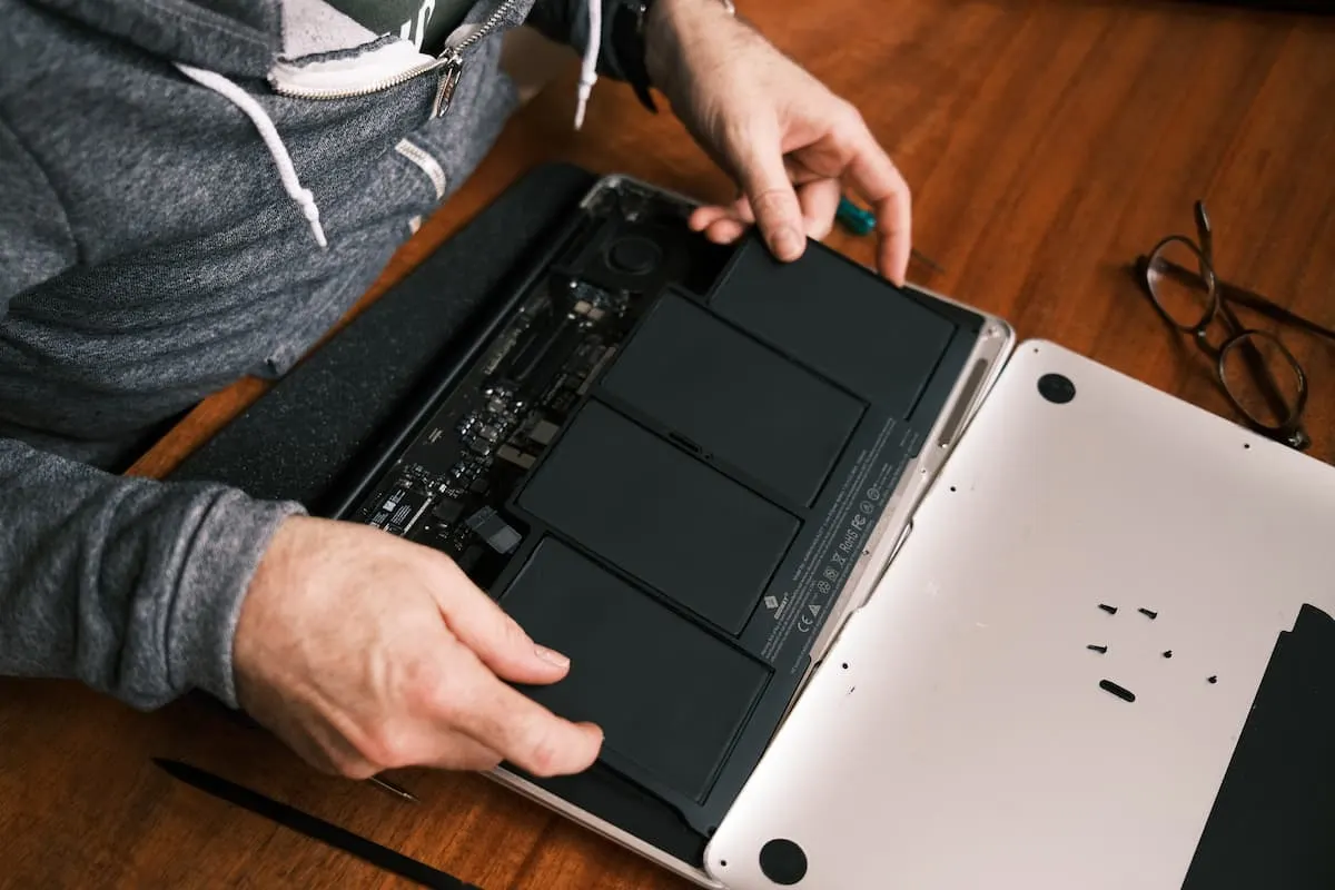 Manos de una persona manipulando la batería interna de un MacBook abierto y desmontado, colocado sobre una mesa de madera.