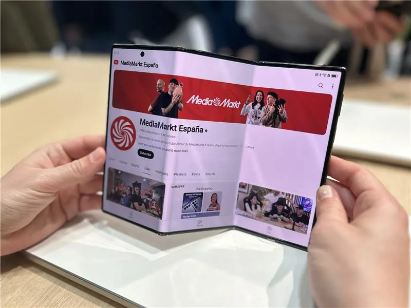 Manos sostienen un teléfono plegable mostrando la página de MediaMarkt España en una mesa.