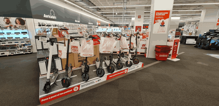 Interior de tienda con fila de patinetes eléctricos en exhibición, bolsas de compra y estanterías con productos electrónicos.