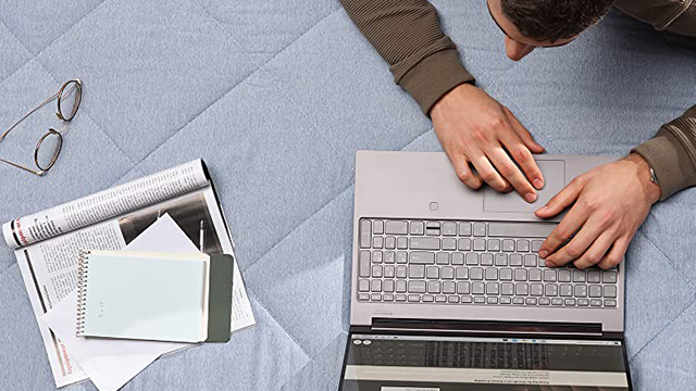 Hombre usando un portátil gris sobre una colcha. Cerca, gafas, revista, bloc de notas y papeles.