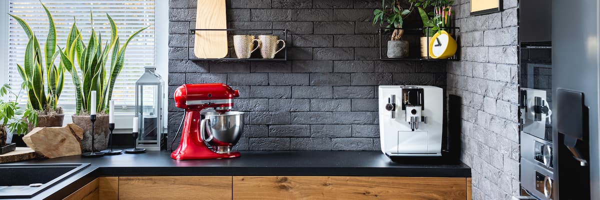 Cocina moderna con pared de ladrillo gris, batidora roja y cafetera blanca sobre encimera de madera con plantas decorativas.