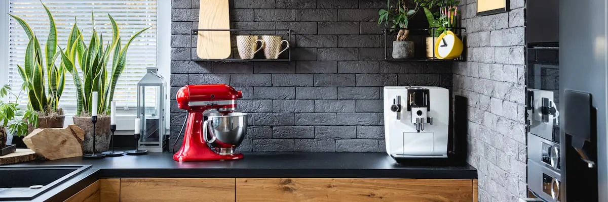 Cocina moderna con pared de ladrillo gris, batidora roja y cafetera blanca sobre encimera de madera con plantas decorativas.