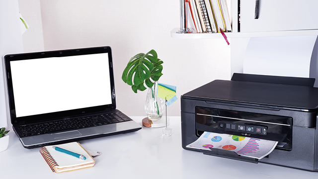 Escritorio blanco con un portátil con pantalla blanca, una impresora imprimiendo, una planta, libreta y estantería con libros.