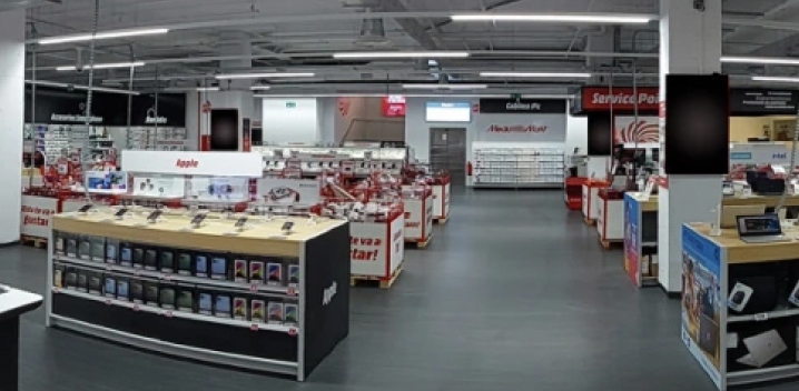 Interior de una tienda con estantes llenos de electrónica y accesorios, bajo luces fluorescentes. Se ven pasillos grises.
