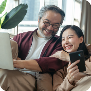 Una pareja sonriente sentada en un sofá usando una laptop y un teléfono móvil en un espacio iluminado con una planta.