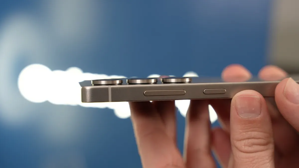 Unas manos sostienen un teléfono gris con tres botones circulares y un botón ovalado en el borde, sobre un fondo azul difuminado.